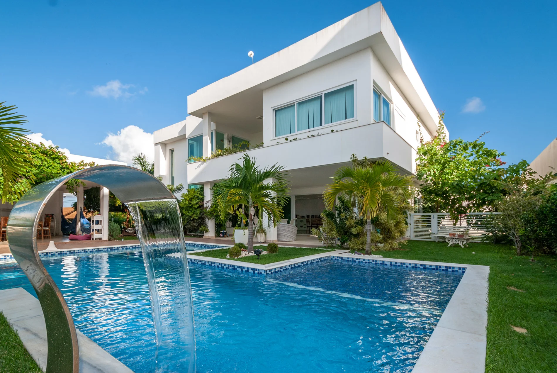 Imagem mostra uma casa de alto padrão branca com uma piscina em foco, cheia de paisagismo ao redor